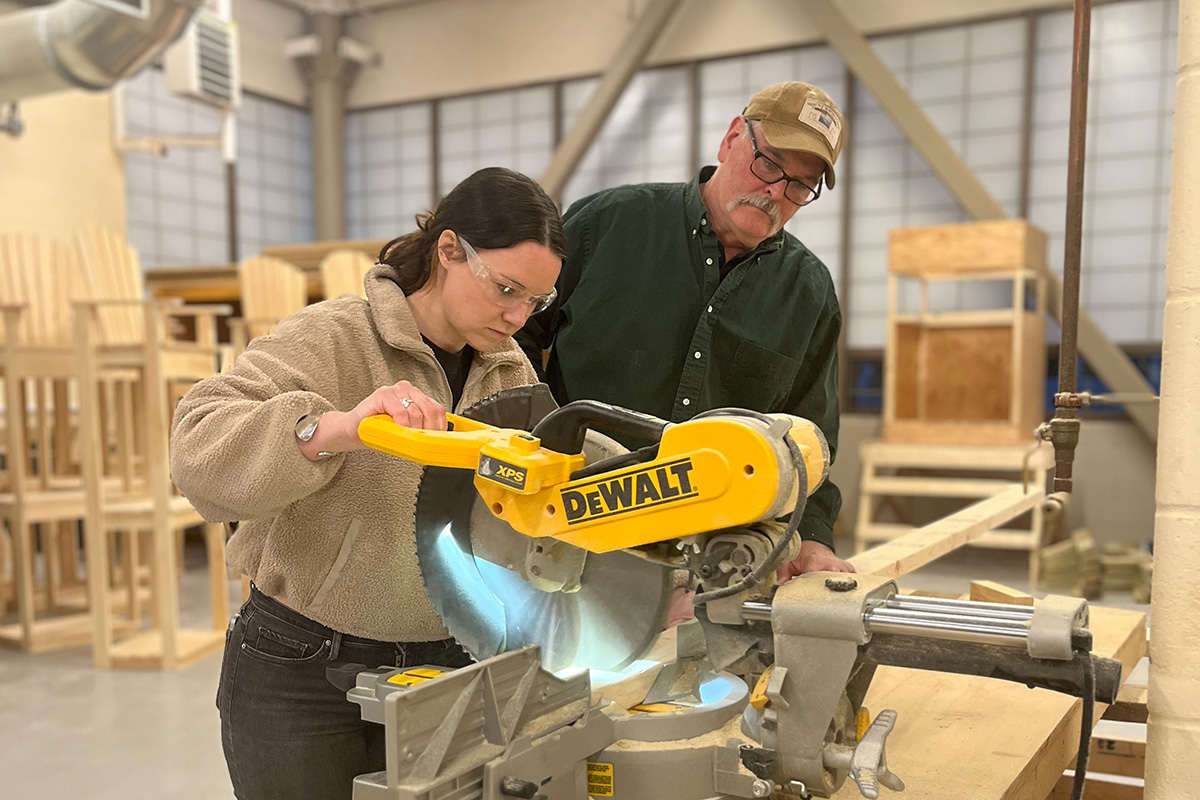 Woman using a chop saw in the carpentry class with instructor looking on
